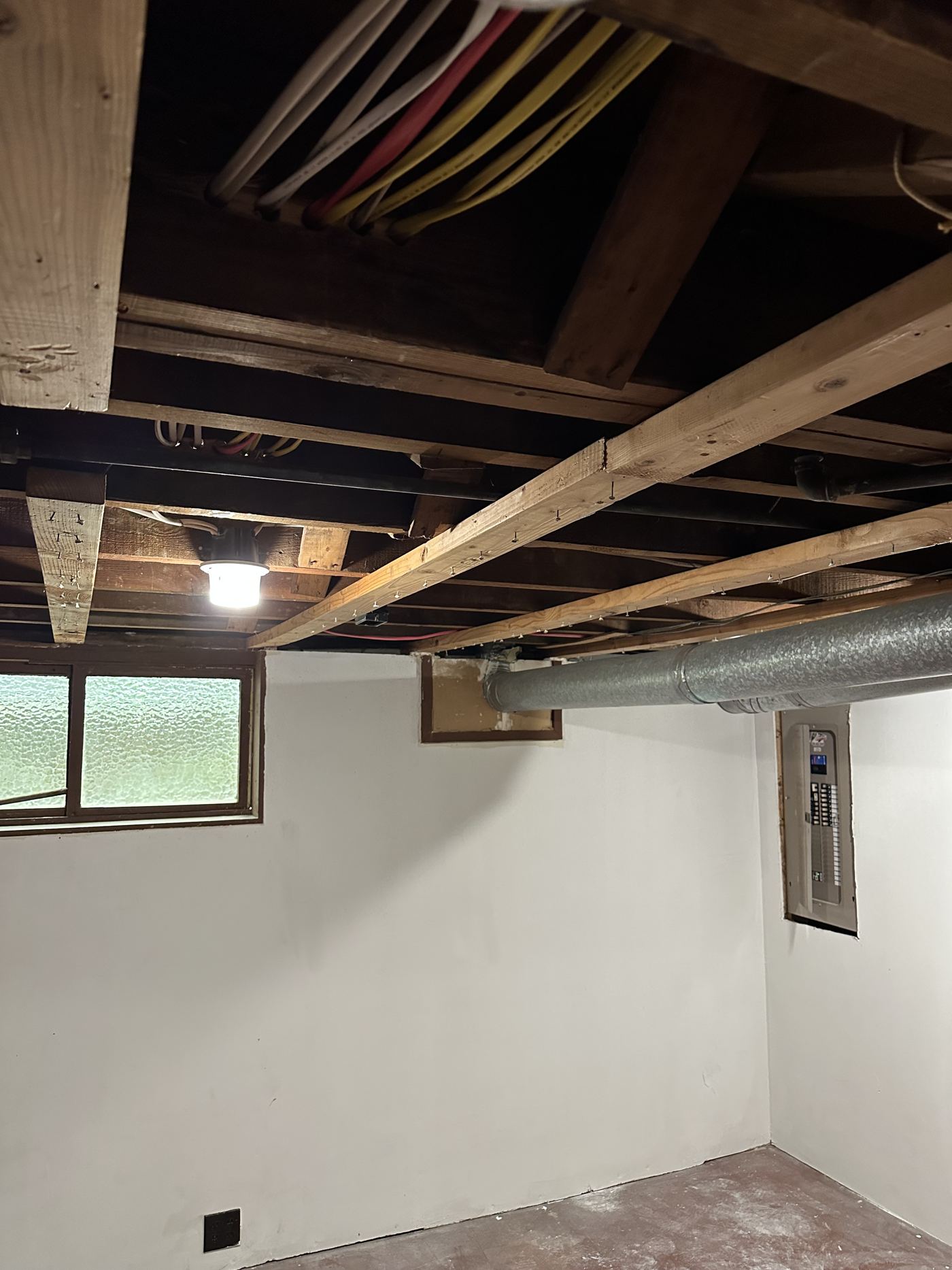 A white-walled basement room with a single silver metal duct running along the ceiling beside the electrical panel