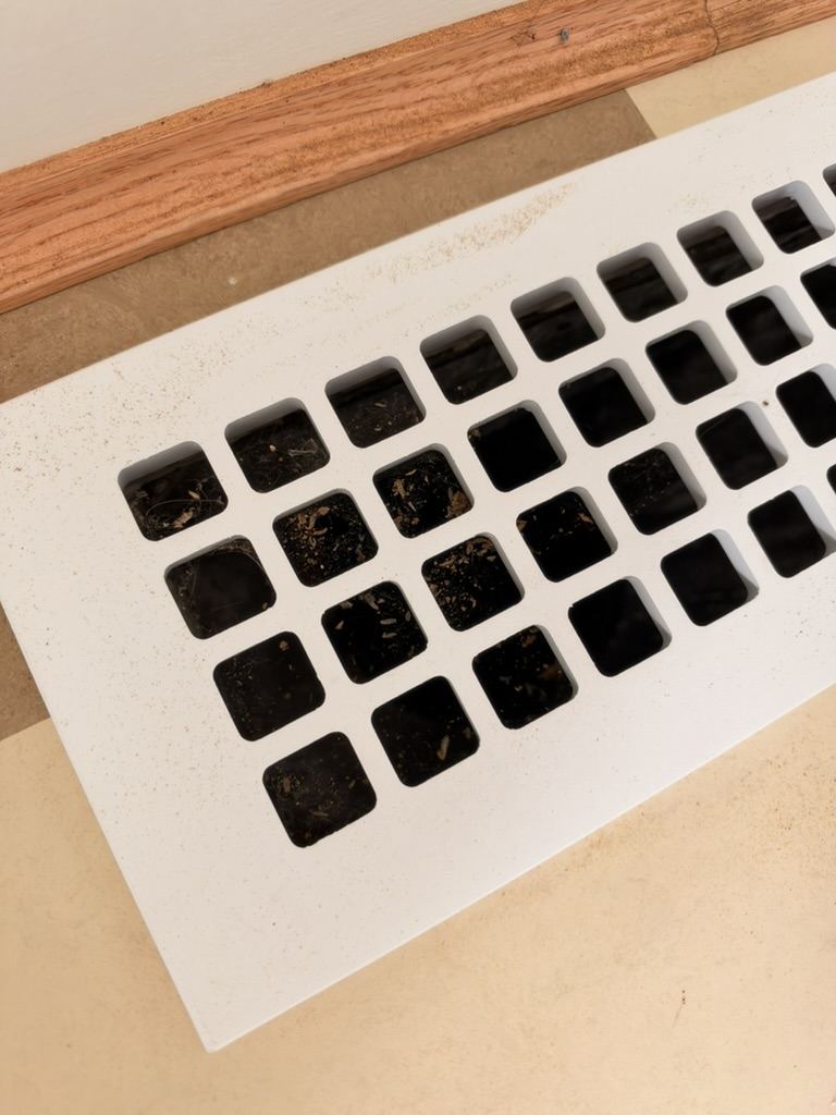 A white floor register grille with dirt and debris caught in and around its openings