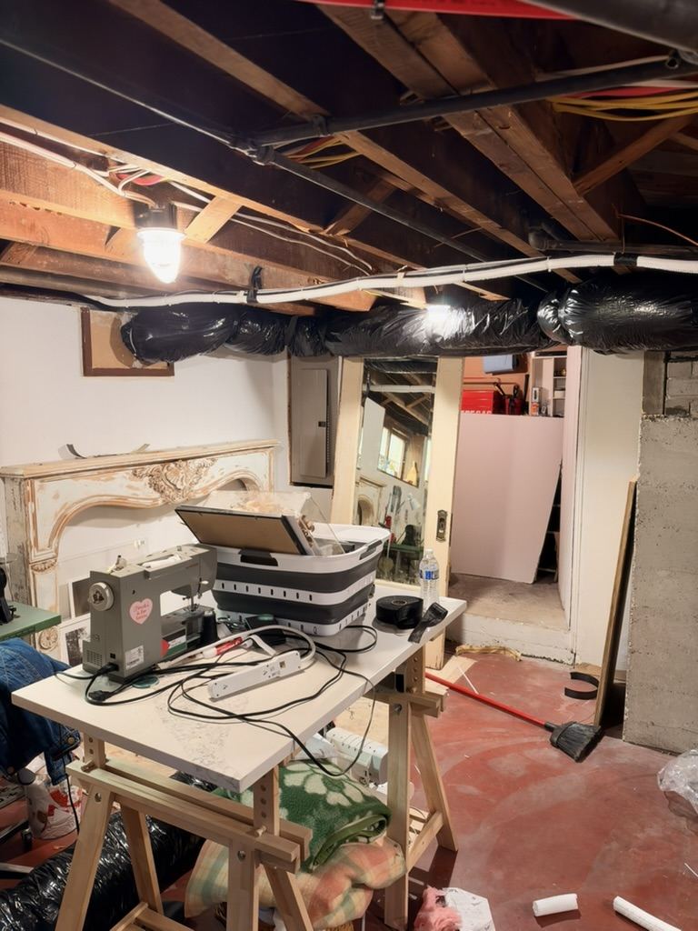 Bulky black flexible duct strung along the basement ceiling joists above a work table