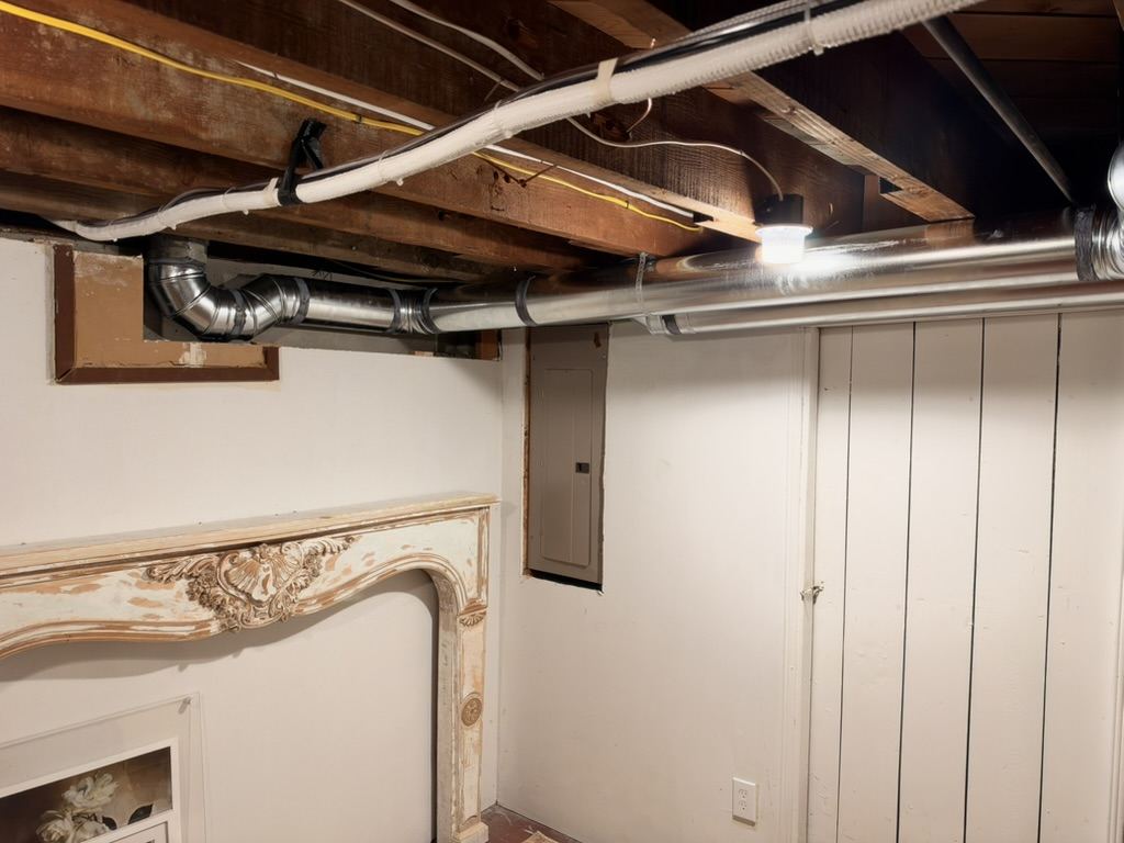 Silver rigid metal duct running along the basement ceiling above the carved mantel, with a white-wrapped refrigerant line set strung along the joists above it